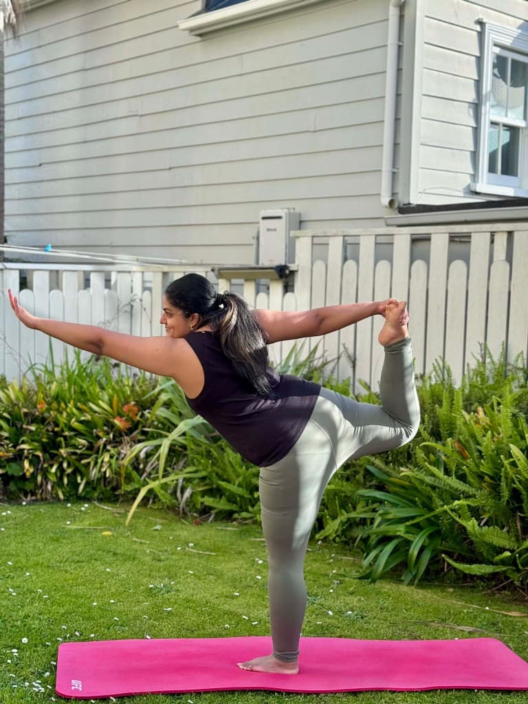 Anu balancing in dancer pose on a mat in a sunlit garden